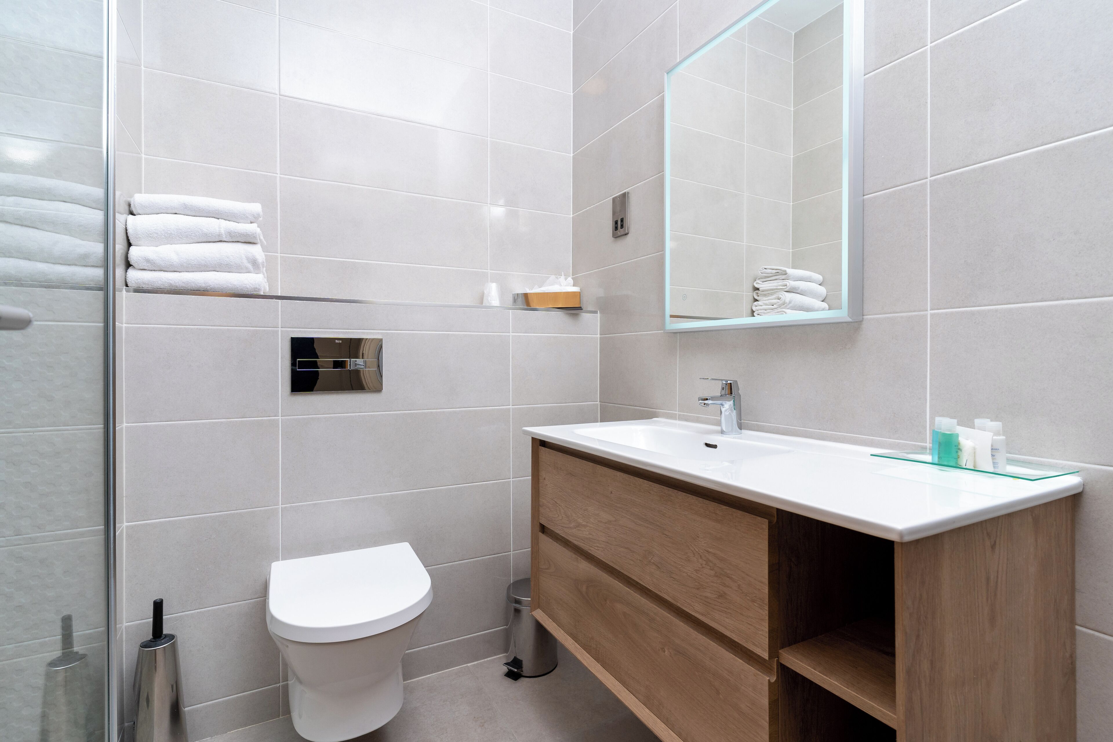 A modern bathroom with a white toilet, a wooden vanity with a mirror, and neatly folded towels on a shelf.