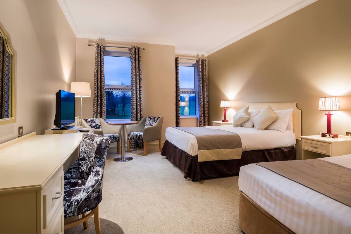 Une chambre d’hôtel avec un lit, un bureau, une chaise, une télévision et deux fenêtres avec des rideaux.