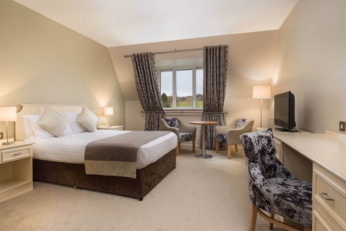 Une chambre d’hôtel avec un grand lit, un bureau avec une télévision, deux chaises, une petite table et des rideaux.