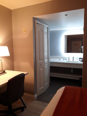 Quarto de hotel com escrivaninha, cadeira e banheiro com pia e espelho.