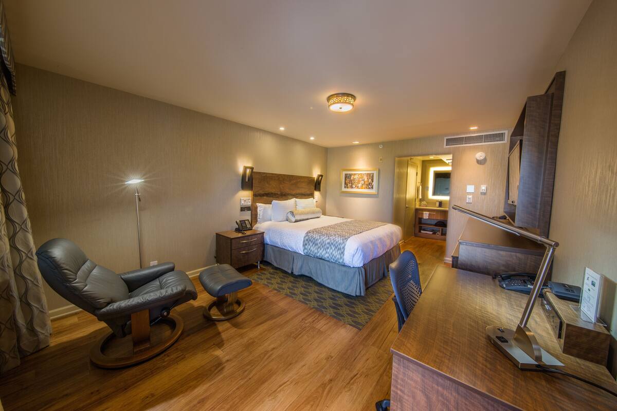 A hotel room with a bed, a desk, a chair, a desk lamp, a bedside table, and a wall-mounted TV.