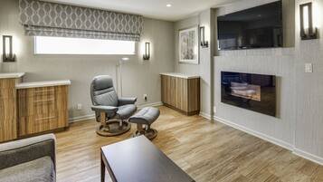 Een moderne woonkamer met een open haard, houten kasten, een grijze fauteuil en een rond poef.