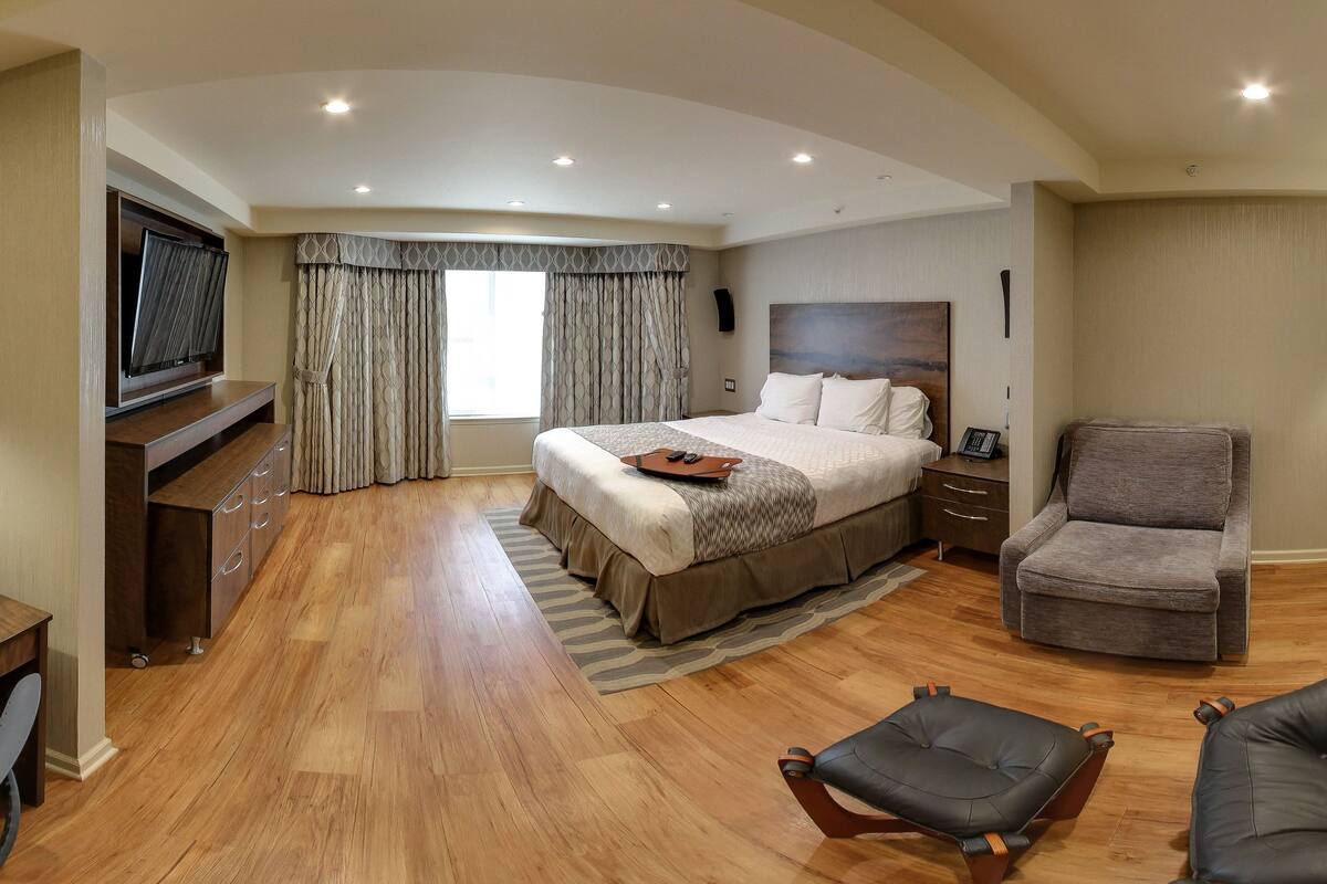 A spacious hotel room with a large bed, a flat-screen TV, a grey armchair, a leather ottoman, and a fireplace.