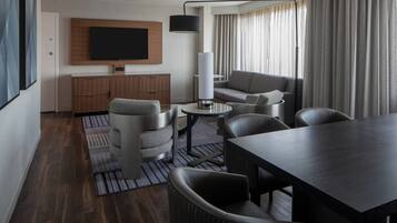 Una habitación de hotel moderna con piso de madera oscura, un sofá gris, una mesa de comedor con sillas, un televisor de pantalla plana montado en la pared y un ventanal con cortinas.