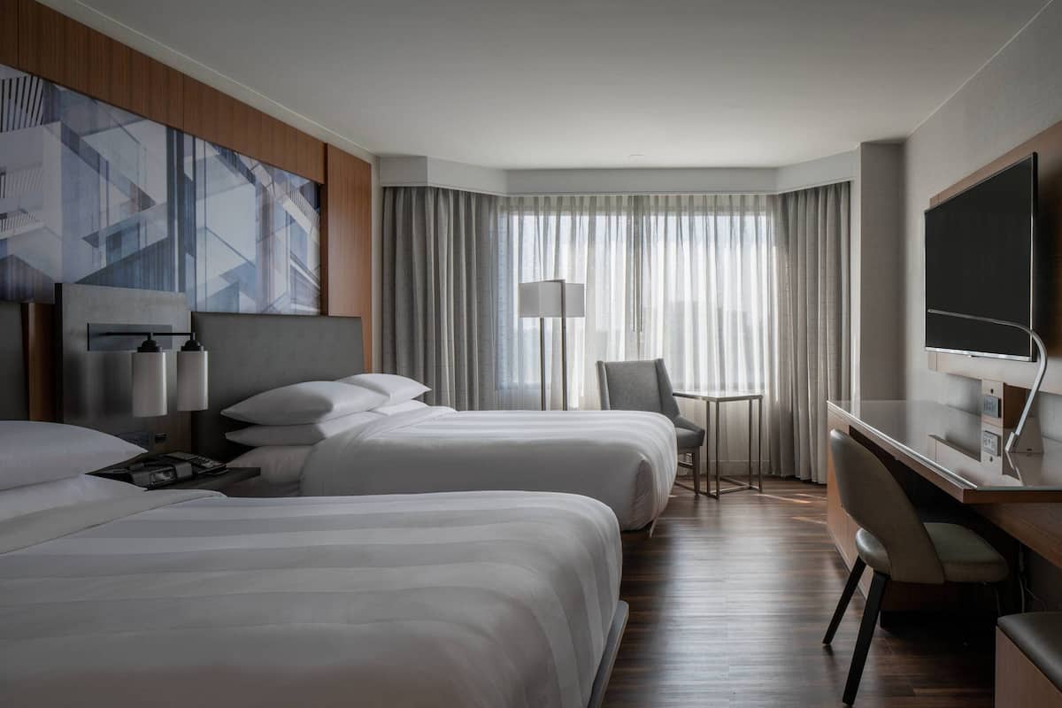 A hotel room with two beds, a desk, a chair, a TV, and a large window with curtains.
