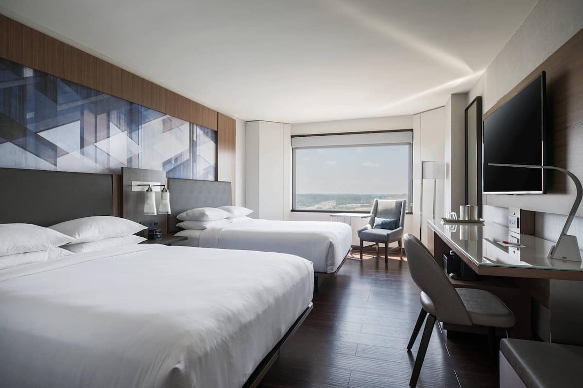 A hotel room with two beds, a desk, a chair, a TV, and a large window offering a view.