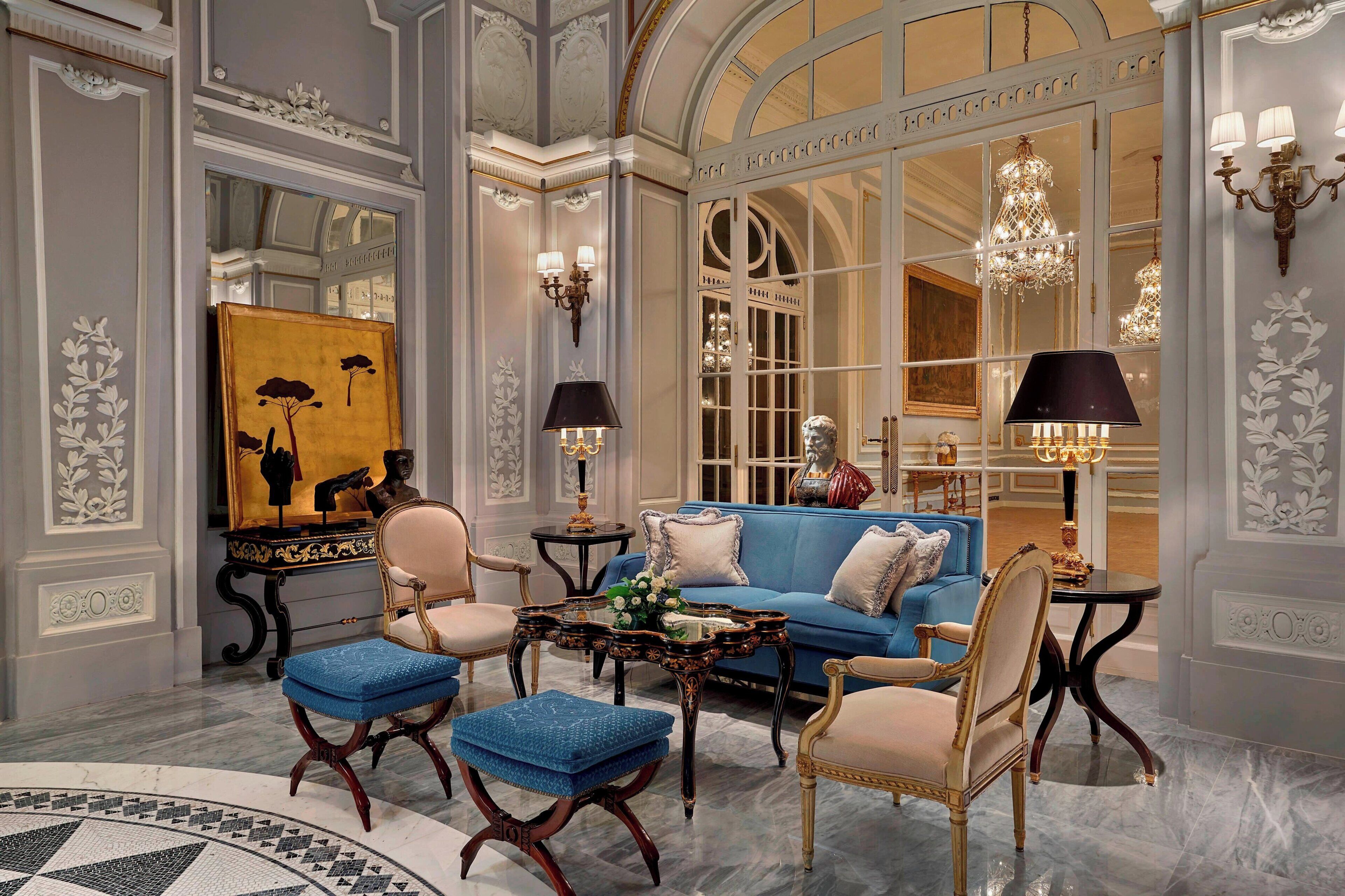 Een luxe kamer met een blauwe bank, bijpassende voetenbankjes, een klassieke fauteuil, een tafeltje met een vaas, en een schilderij aan de muur.