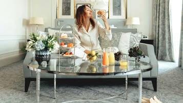 A woman sitting on a grey sofa in a well-lit room with a glass coffee table, a vase with flowers, and a tray with food and drinks.