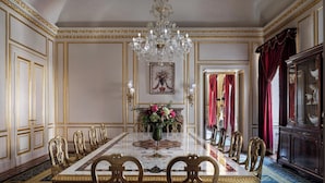 A formal dining room with a large marble table, ornate chairs, a chandelier, and a red curtain.
