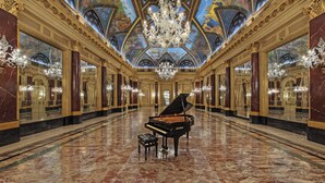 Un gran salón con un piano de cola negro, candelabros ornamentales y un techo con un fresco detallado.