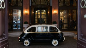 A classic black car parked in front of a building with a grand entrance, large windows, and decorative wall sconces.
