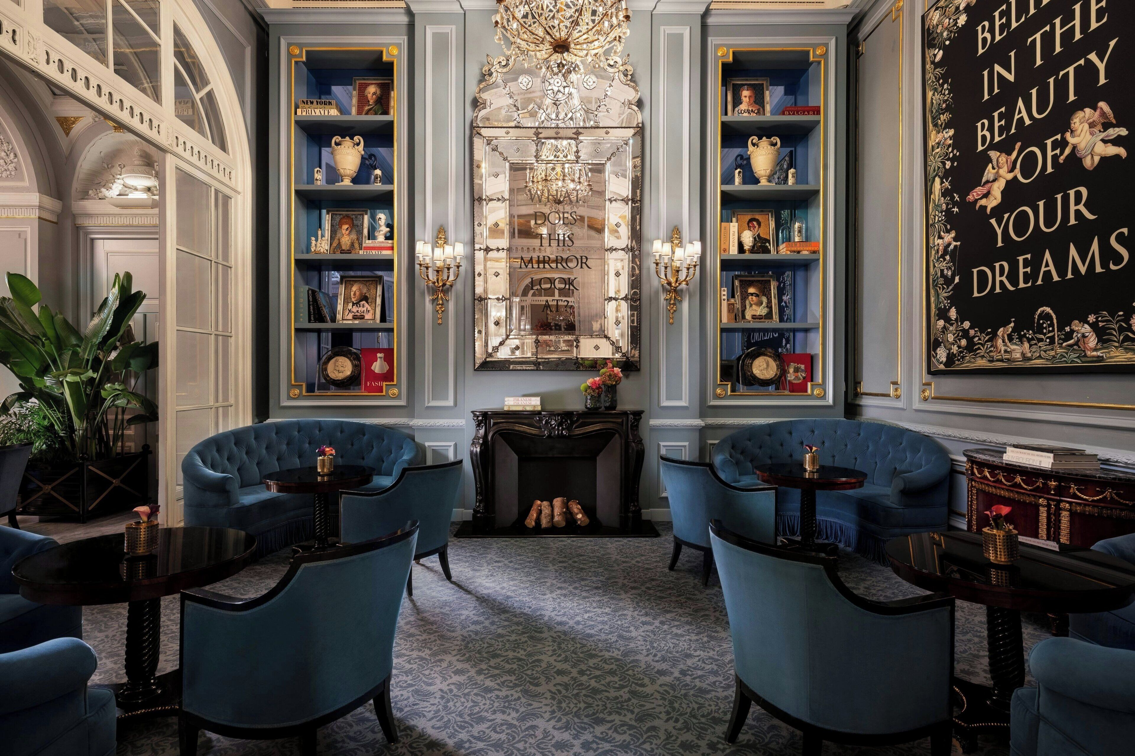A luxurious room with a fireplace, blue armchairs, a bookshelf, and a chandelier.