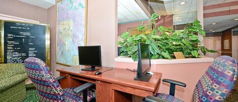 A hotel reception area with a wooden desk, two patterned chairs, a computer monitor, a large map on the wall, and a potted plant.