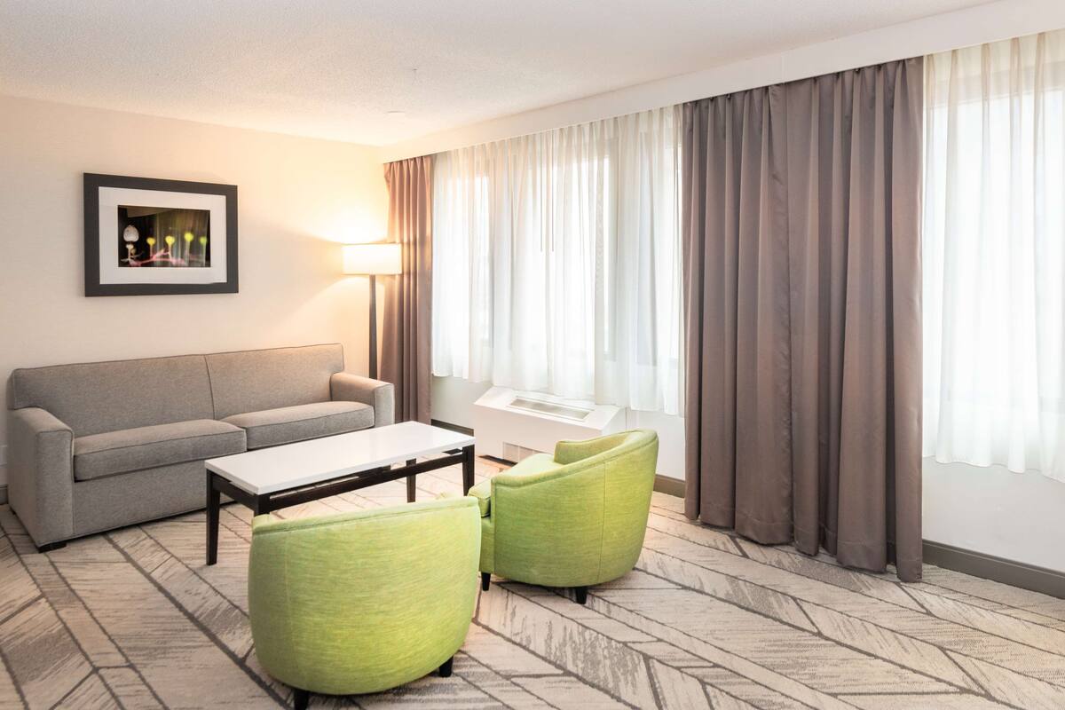 A hotel room with a grey sofa, a white coffee table, two green armchairs, a framed picture on the wall, and a large window with curtains.
