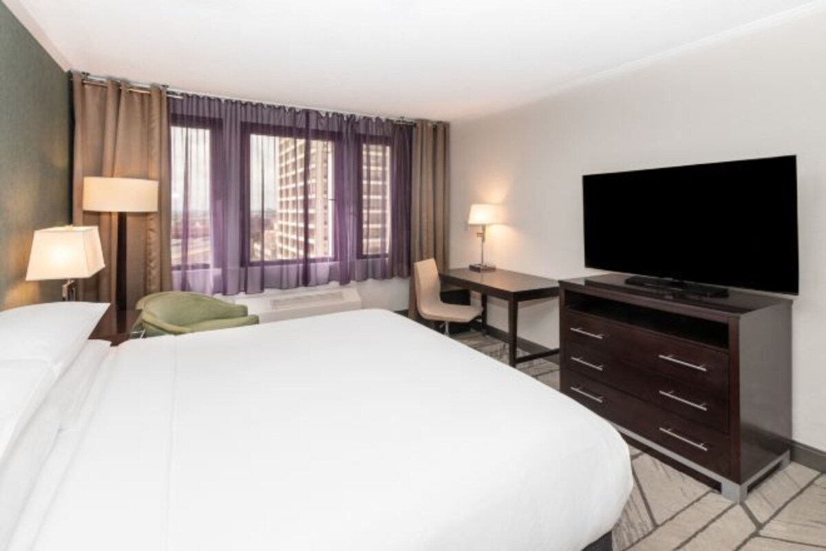 A hotel room with a large bed, a desk with a chair, a television, and a window with curtains.