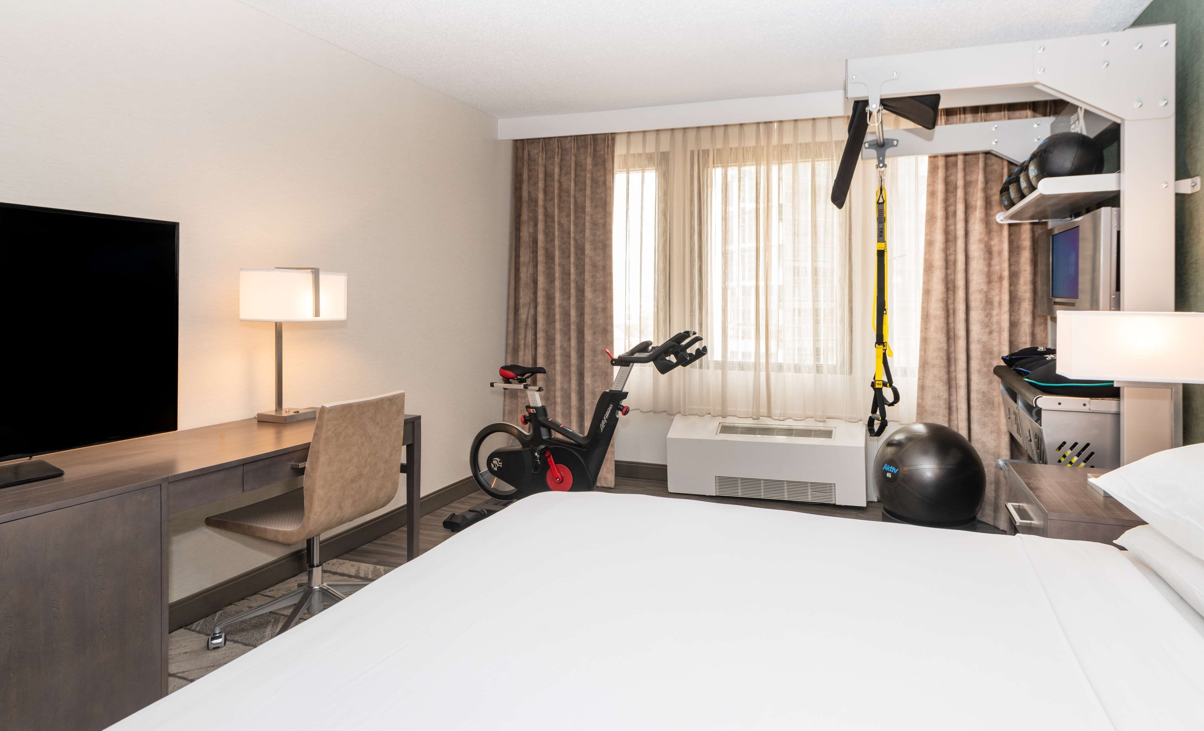 Habitación de hotel con cama, escritorio con televisor, silla, bicicleta estática y cinta de correr.