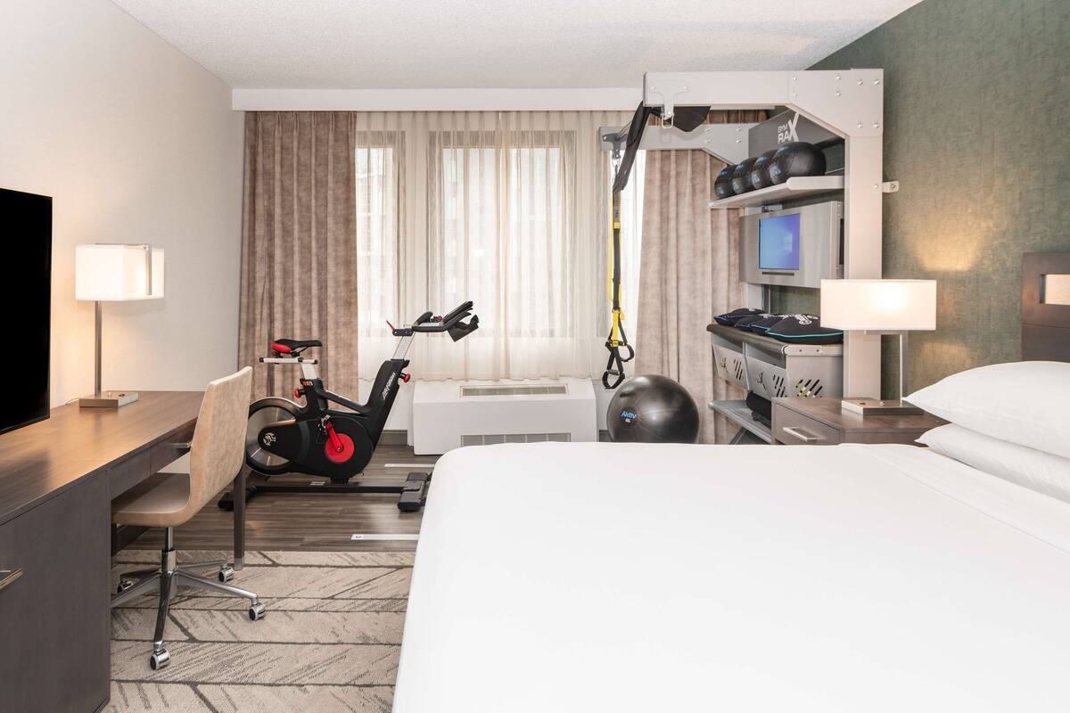 A hotel room with a bed, a desk with a flat-screen TV, a treadmill, a stationary bike, and a massage chair.
