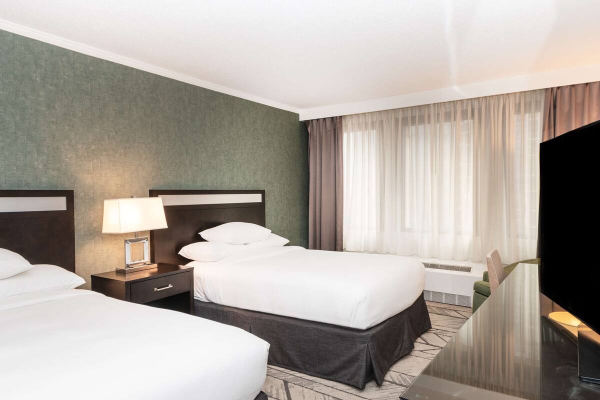 A hotel room with two beds, a flat-screen TV, a bedside table with a lamp, and a large window with curtains.