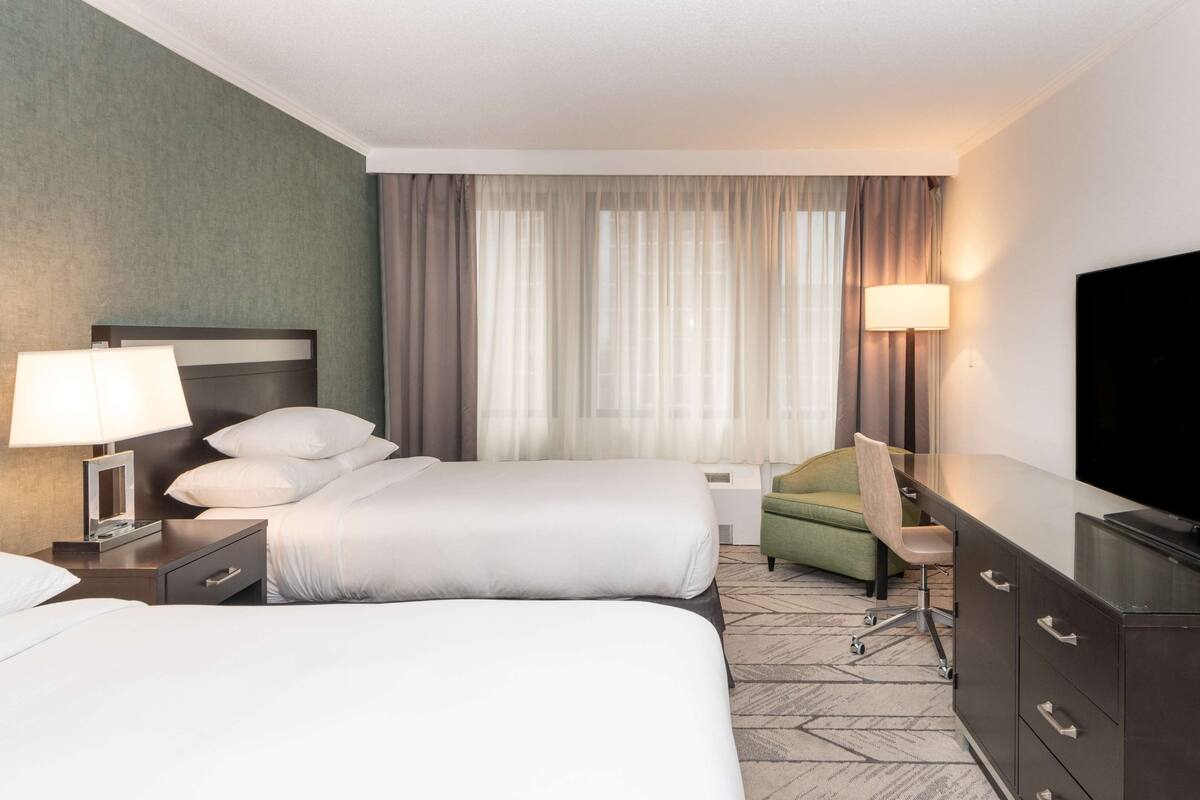 A hotel room with two beds, a desk, a chair, a TV, and a lamp.