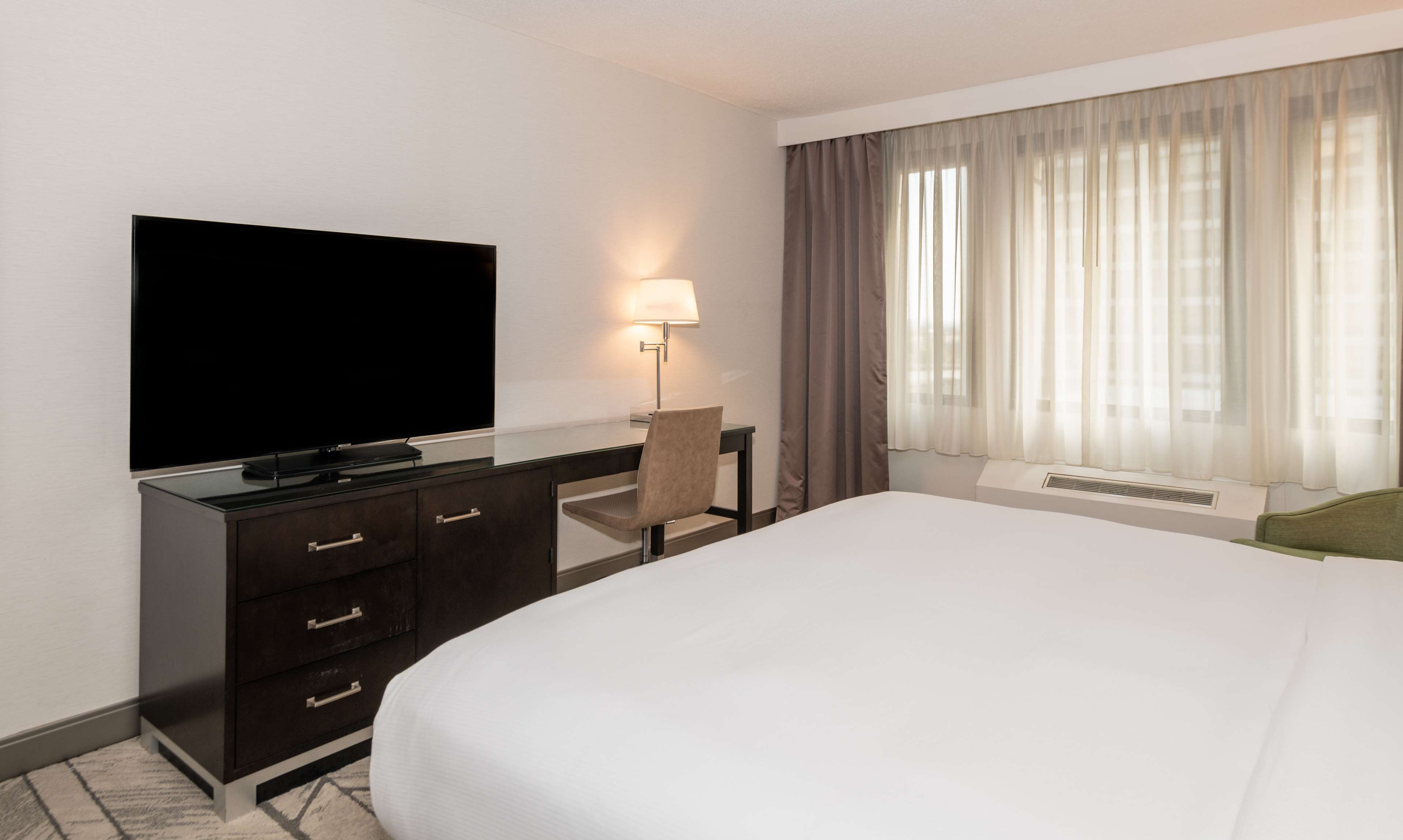 A hotel room with a large bed, a flat-screen TV mounted on the wall, a desk with a chair, and a window with sheer curtains.