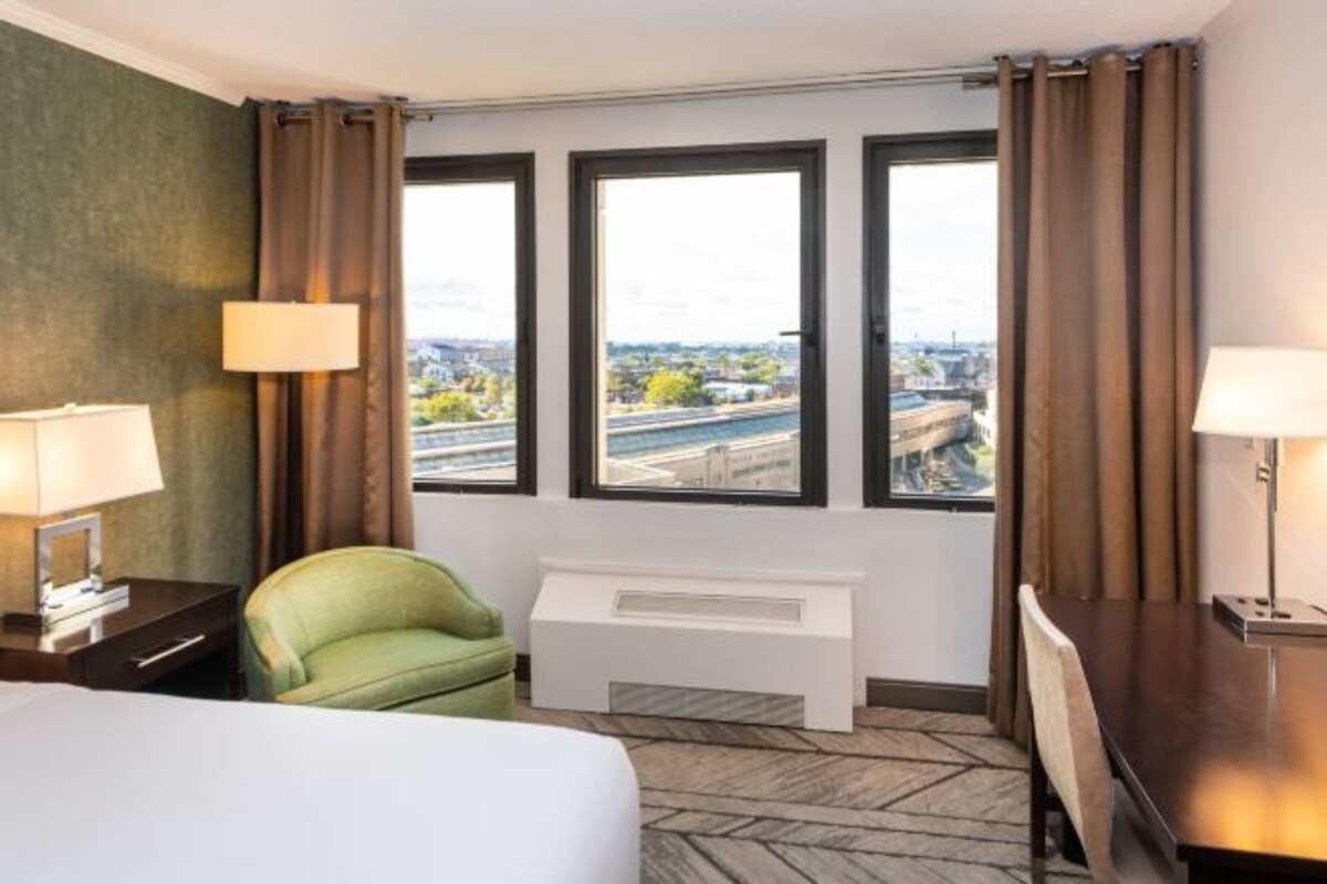 A hotel room with a bed, a green armchair, a desk, and a view of a cityscape through three windows.
