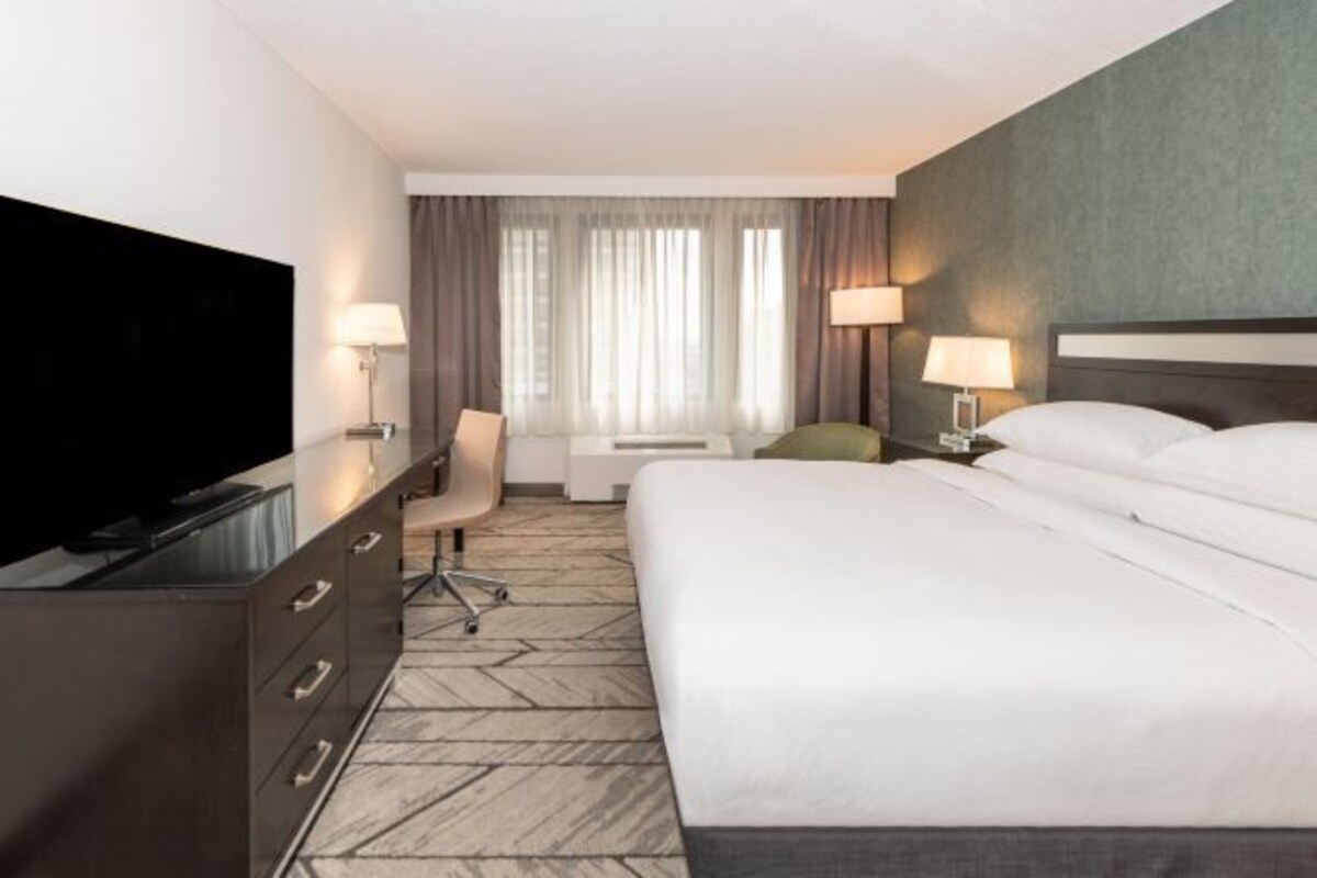 A hotel room with a bed, a desk with a television, a chair, a lamp, and a window with curtains.