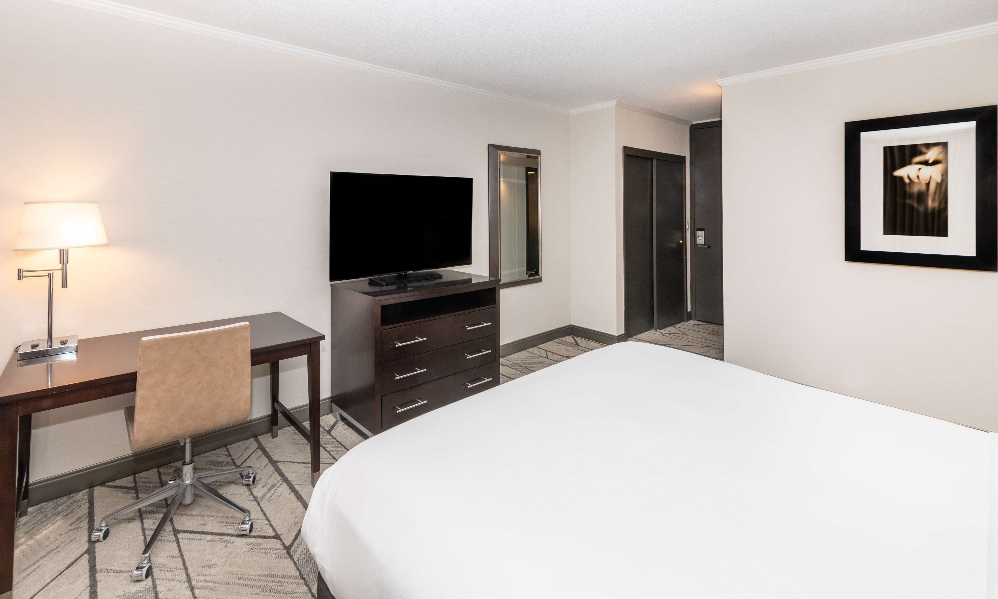 A hotel room with a bed, a desk with a lamp, a TV, a dresser, and a door.