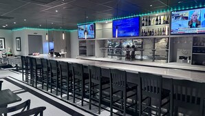 Zona de bar con varias pantallas mostrando noticias, una barra y asientos con taburetes altos.