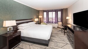 Habitación de hotel con una cama grande, dos mesitas de noche con lámparas, un escritorio con silla, un televisor de pantalla plana y un amplio ventanal con cortinas.