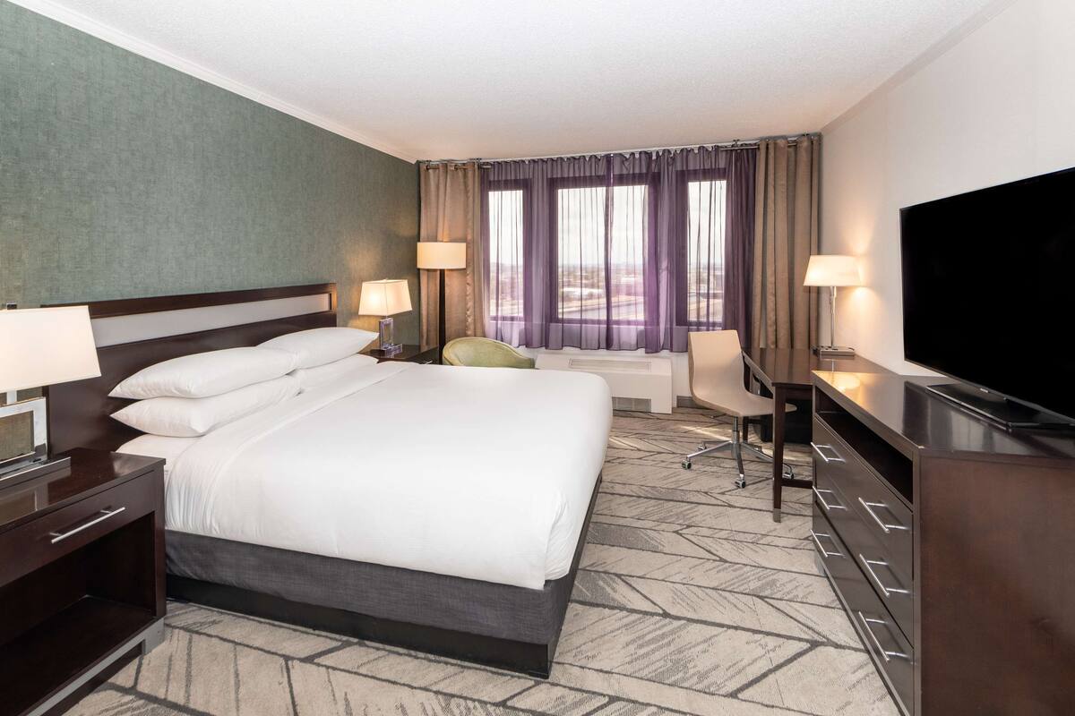 A hotel room with a large bed, two bedside tables with lamps, a desk with a chair, a flat-screen TV, and a large window with curtains.