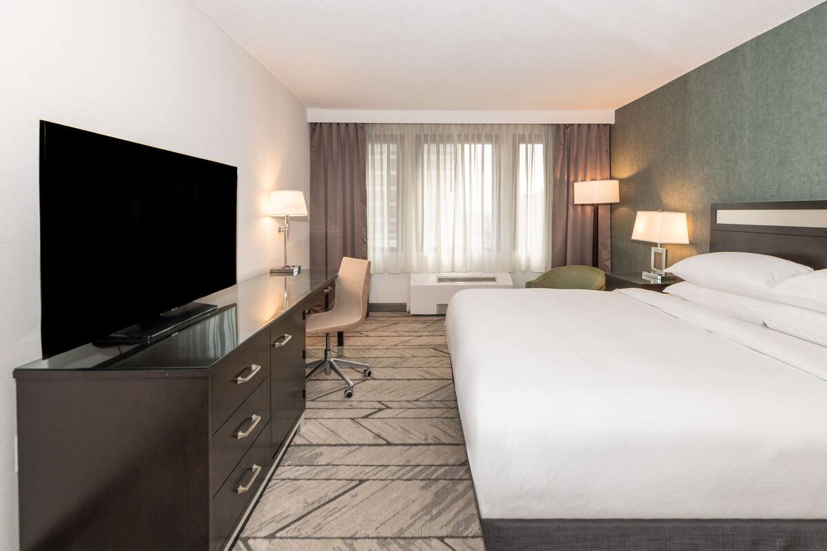 A hotel room with a bed, a desk with a flat-screen TV, a chair, a desk lamp, and a window with curtains.