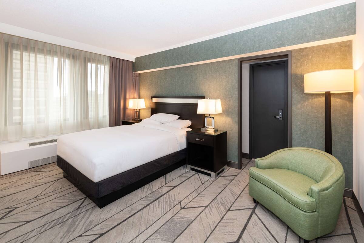 A hotel room with a bed, two bedside tables with lamps, a green armchair, and a door.