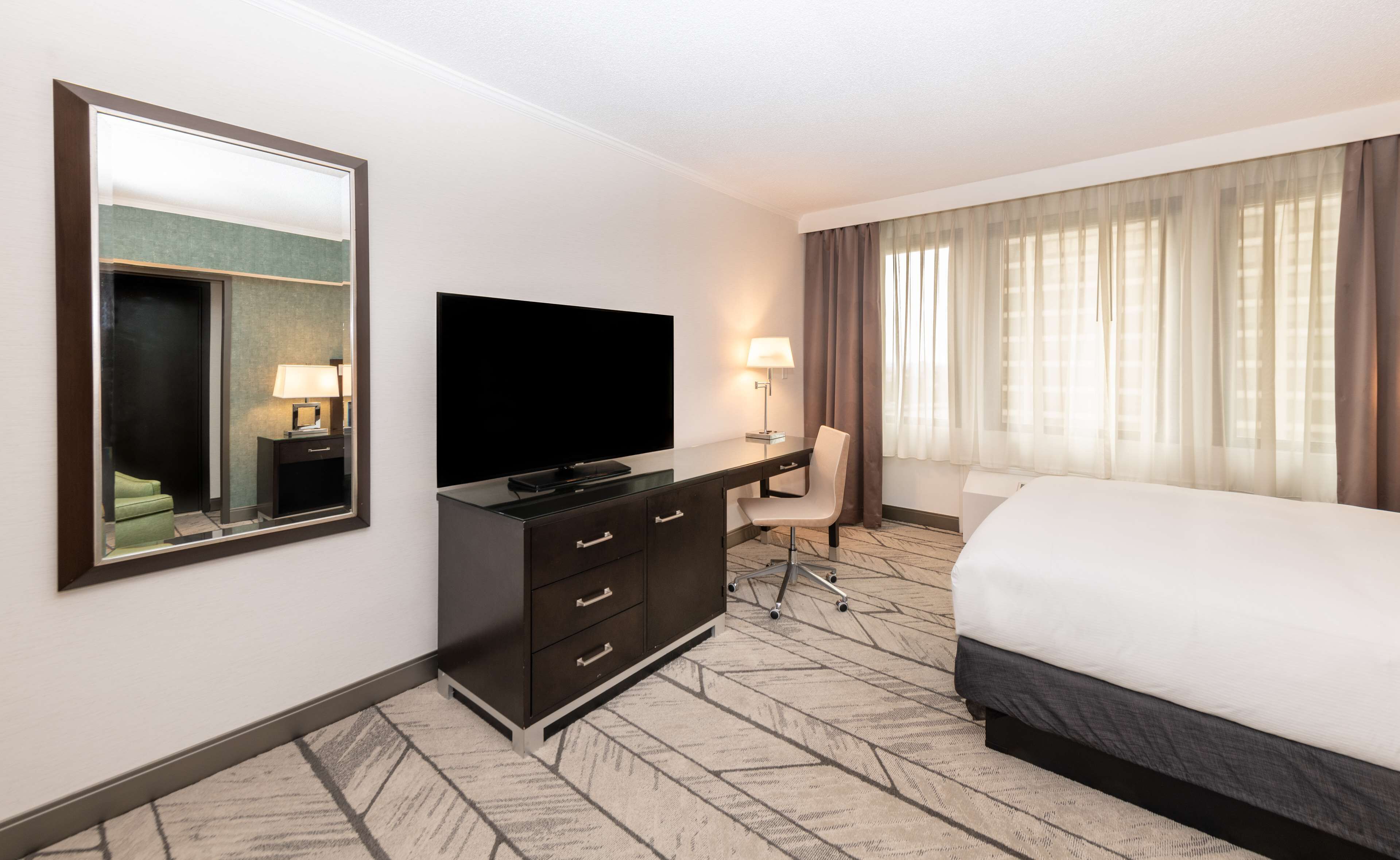 Quarto de hotel com uma cama grande, televisão de tela plana, escrivaninha com cadeira, espelho e janela com cortinas.
