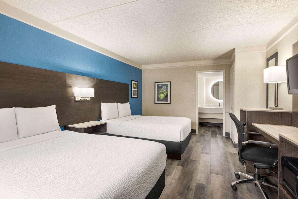 A hotel room with two beds, a desk, a chair, a TV, and a bathroom with a mirror and sink.