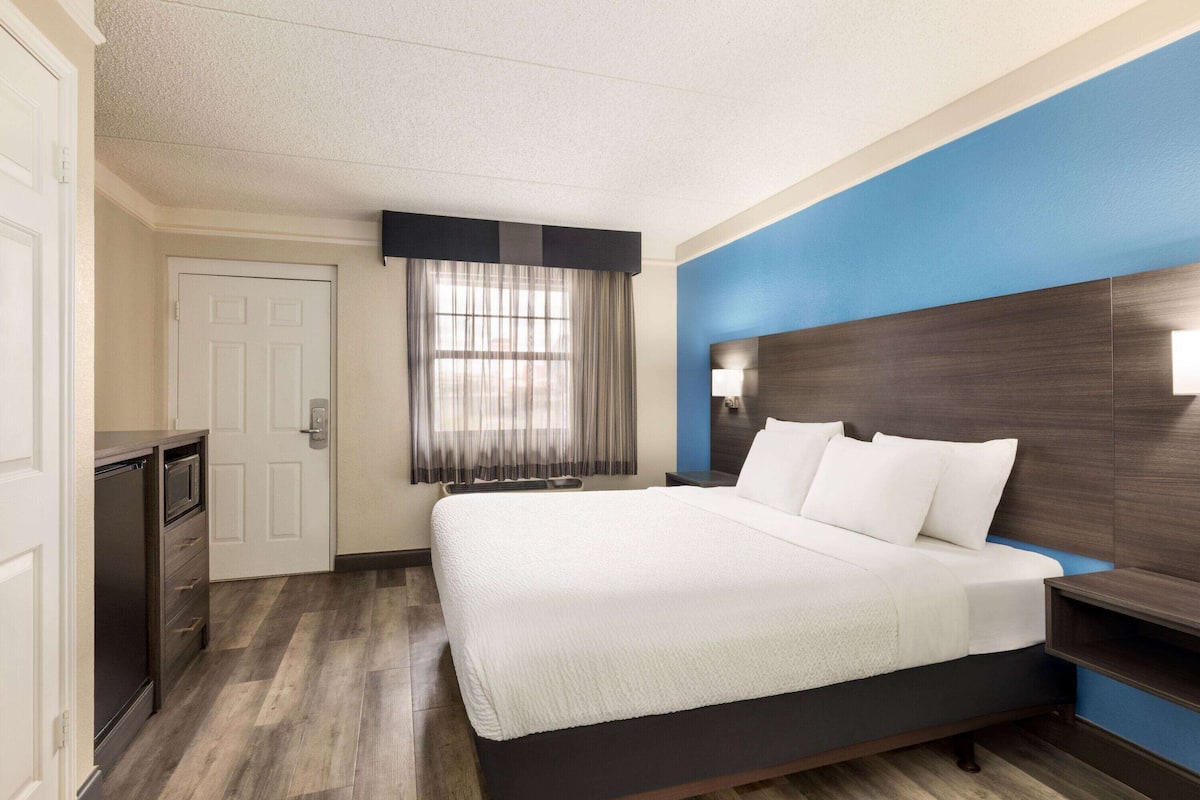 A hotel room with a large bed, a bedside table, a lamp, a window with curtains, a door, and a built-in shelf.