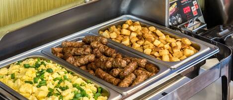 A buffet with three trays of food: scrambled eggs, sausages, and roasted potatoes.
