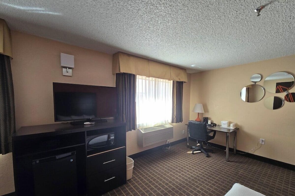 A hotel room with a flat-screen TV, a desk with a chair, a bed, a small table with a lamp, and a wall-mounted mirror.
