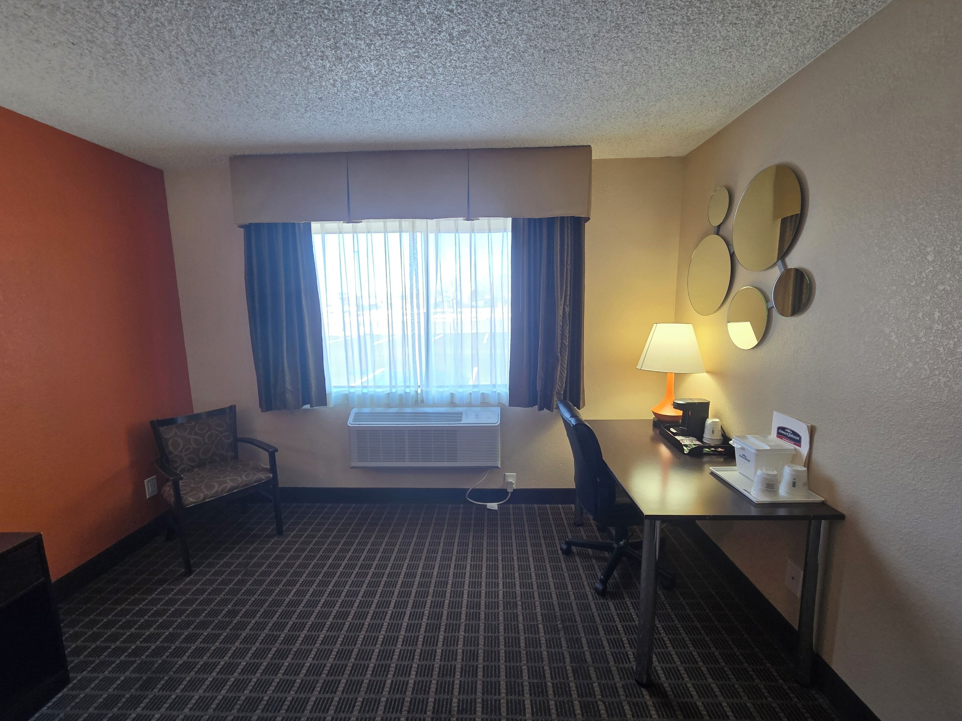 Habitación de hotel con cama, escritorio, silla, ventana con cortinas, espejo montado en la pared y lámpara.