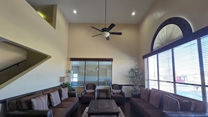 A spacious living room with a high ceiling, a ceiling fan, and large windows with blinds.