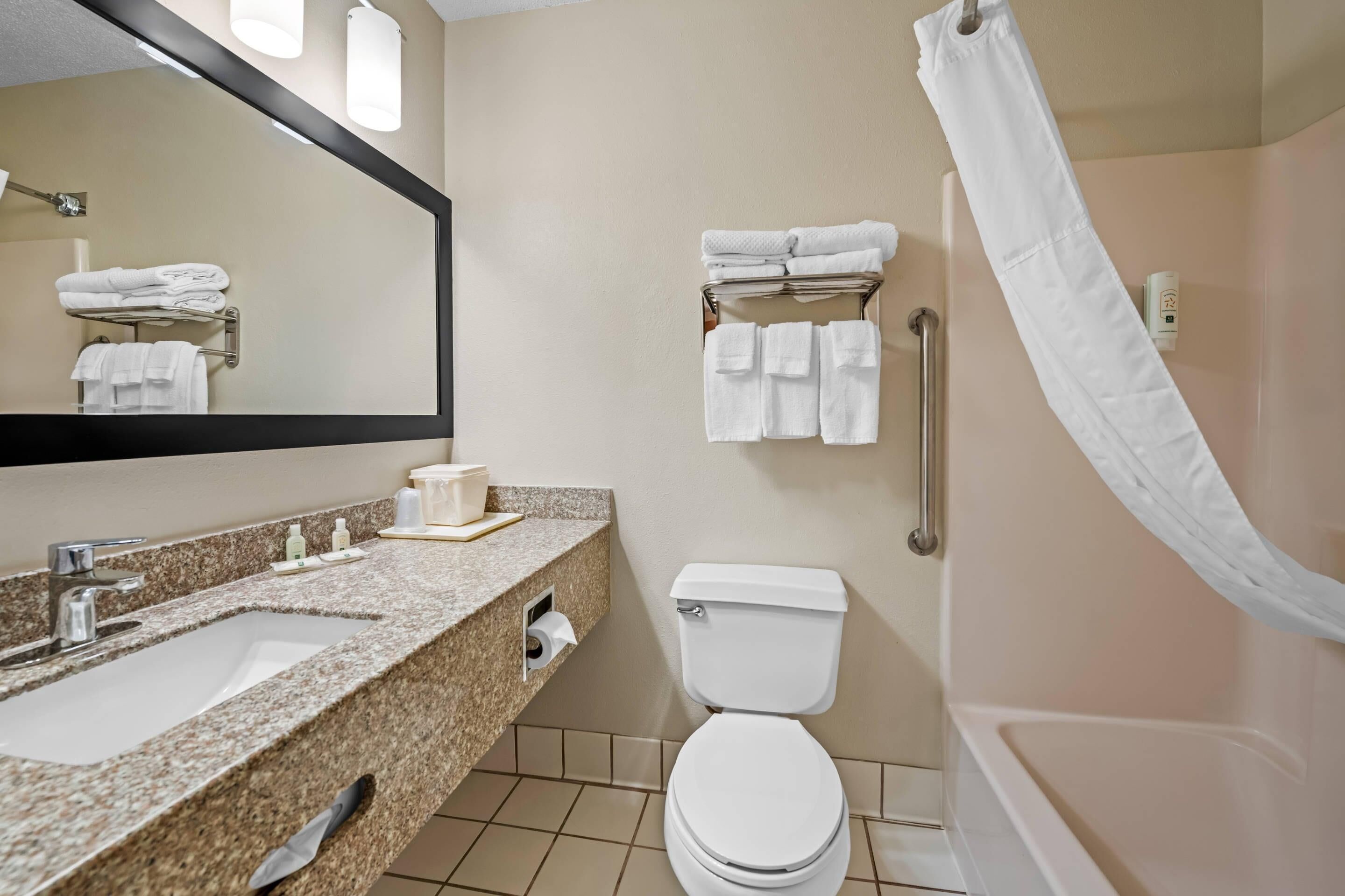 A bathroom with a granite countertop sink, a toilet, a shower with a white curtain, and a towel rack with folded towels.