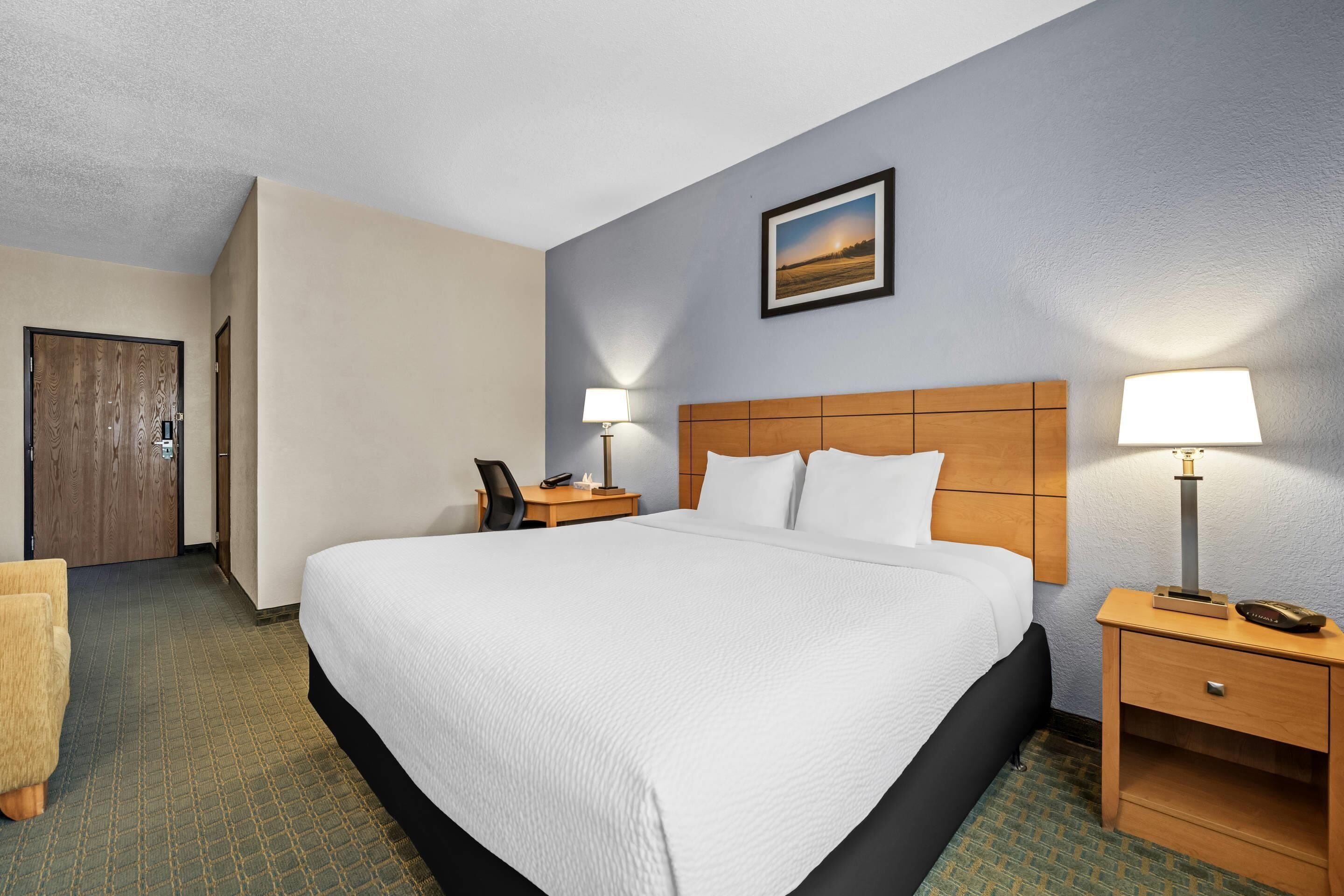 A hotel room with a large bed, two bedside tables with lamps, a wooden headboard, a framed picture on the wall, and a carpeted floor.