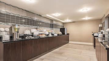 A modern hotel kitchen with a buffet setup, including a coffee machine, fruit bowls, and a refrigerator.