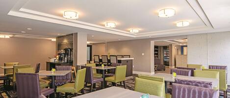 A hotel dining area with multiple tables and chairs, a bar area, and a visible kitchen in the background.
