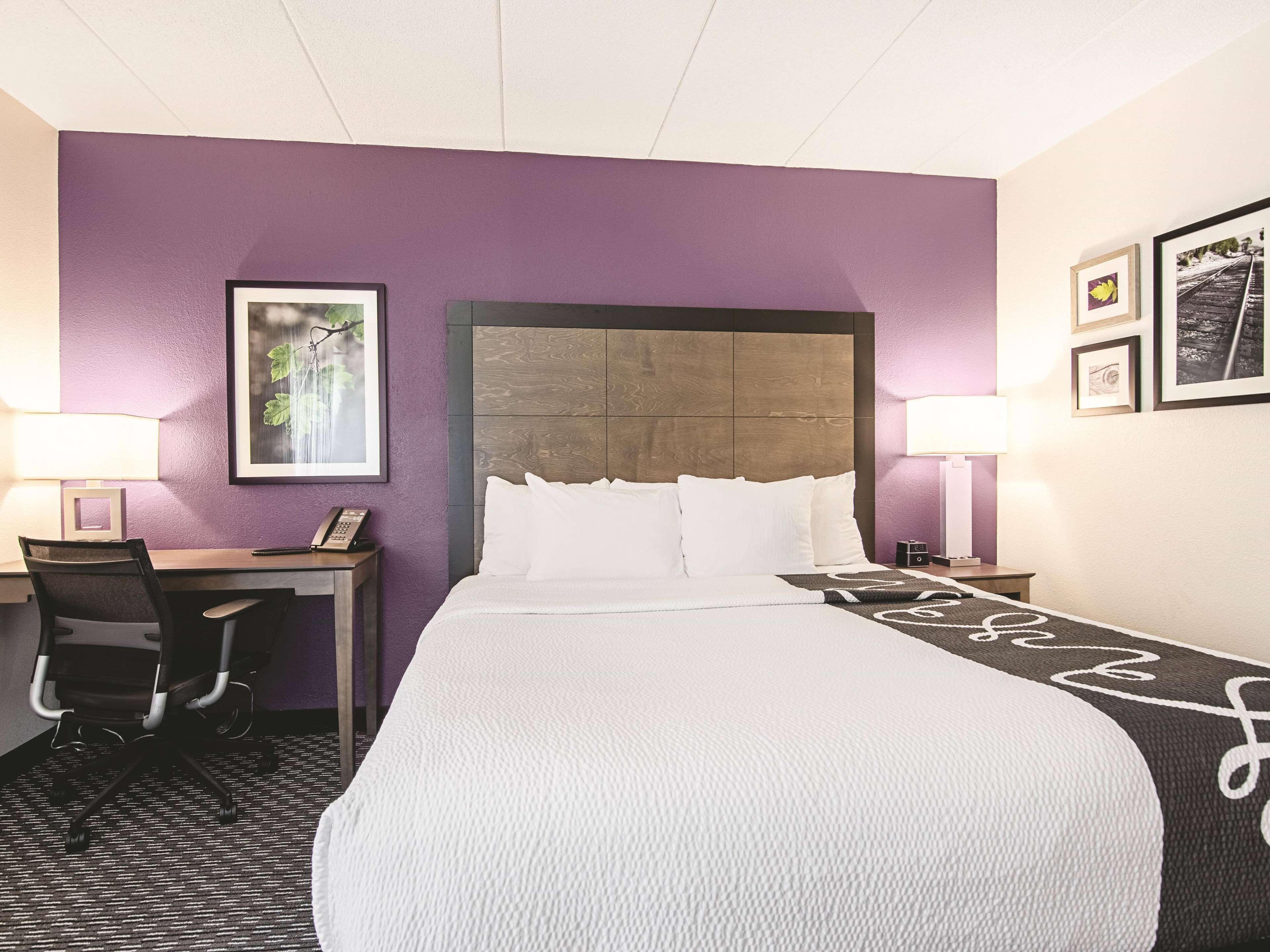 A hotel room with a large bed, a desk, and a chair. There are two bedside lamps and a framed picture on the wall.