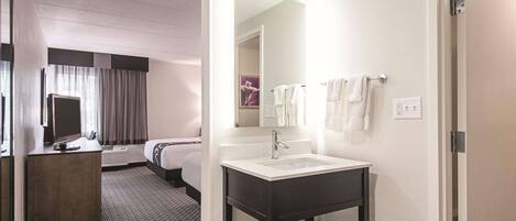 A hotel room with a large bed, a desk with a flat-screen TV, a bathroom with a sink and mirror, and a door leading to another room.