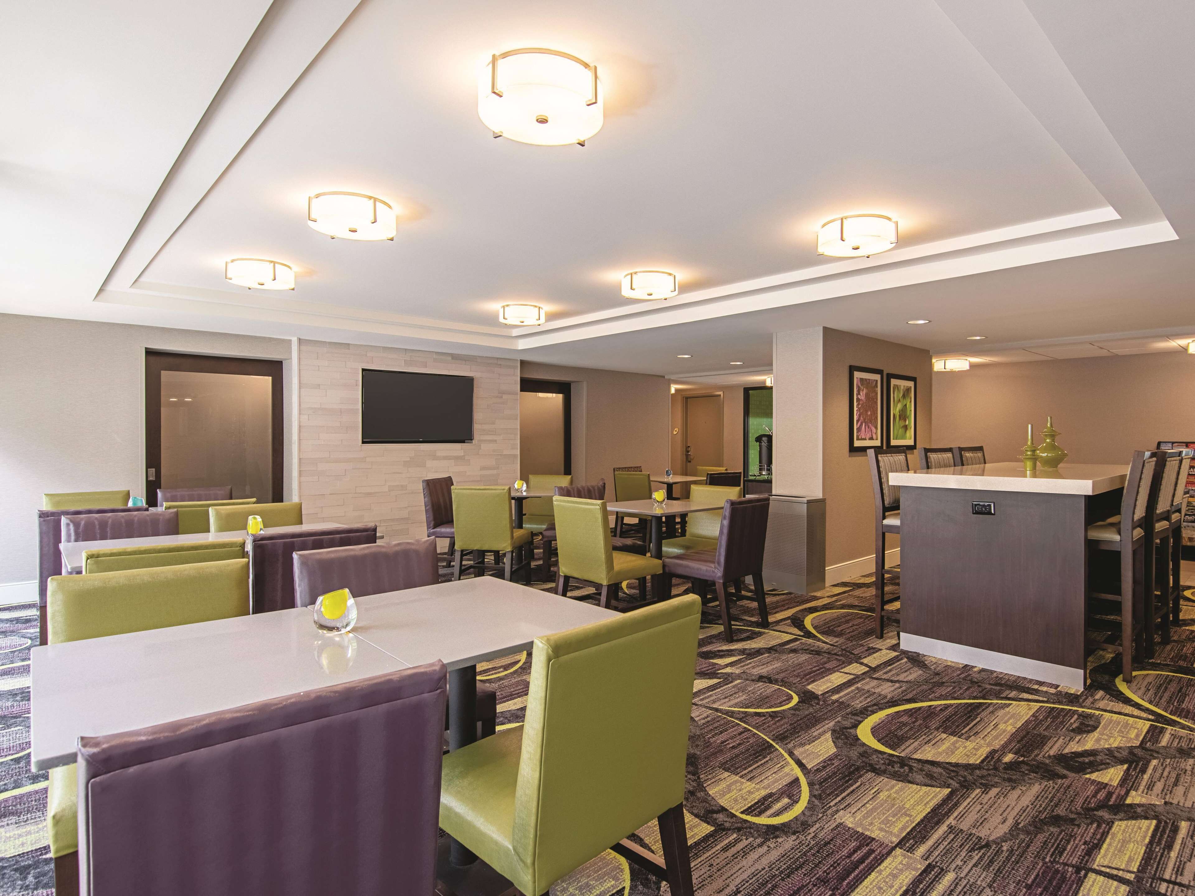 Habitación de hotel con zona de comedor, varias mesas y sillas, un televisor de pantalla plana grande y un minibar.