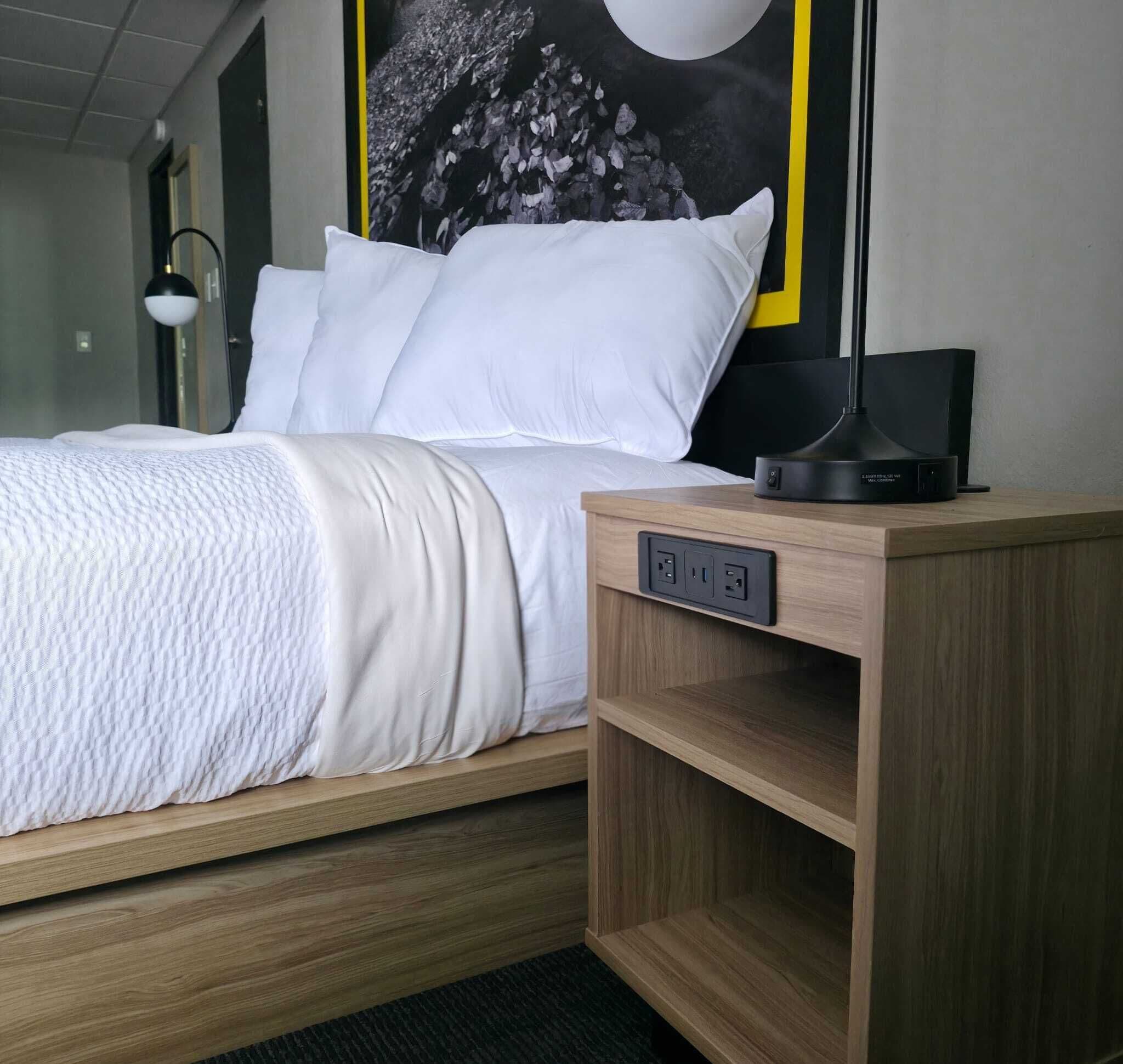 A hotel room with a bed, a bedside table with a lamp, and a wall-mounted power outlet.