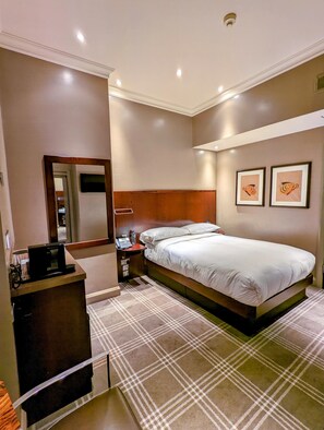 A hotel room with a large bed, a bedside table with a phone and water bottle, a mirror, and two framed pictures on the wall.
