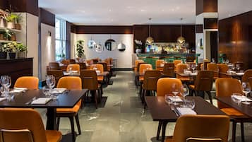 Un restaurante con varias mesas y sillas, una zona de bar y plantas en macetas sobre estanterías.