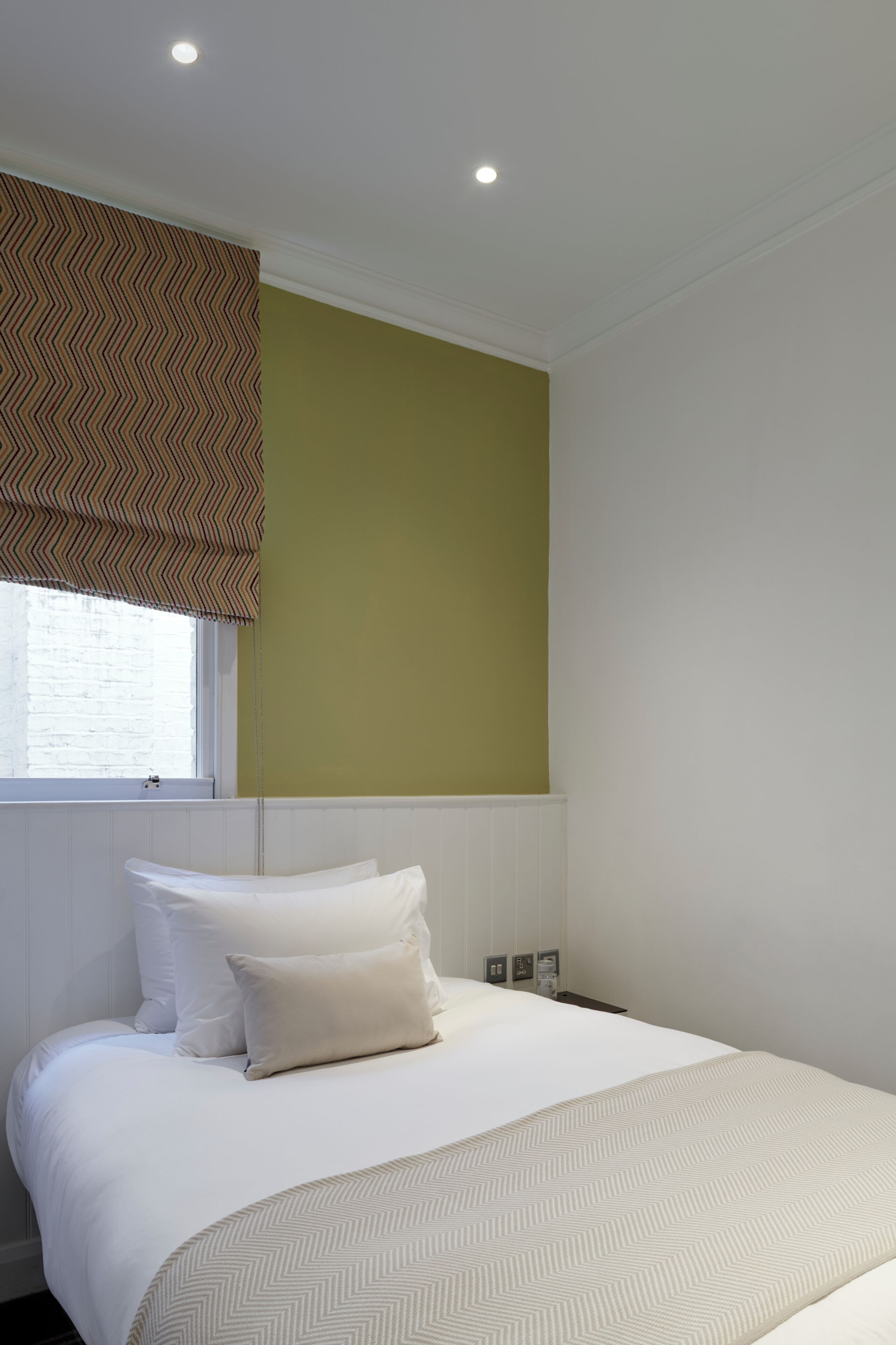 A hotel room with a bed, a window with blinds, a wall with a patterned curtain, and a light switch on the wall.